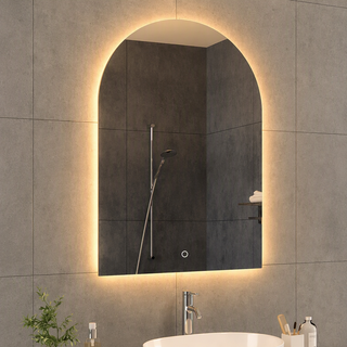 An arched LED-backlit bathroom mirror mounted on a gray tiled wall above a white sink, reflecting a dark shower area with a handheld shower head.
